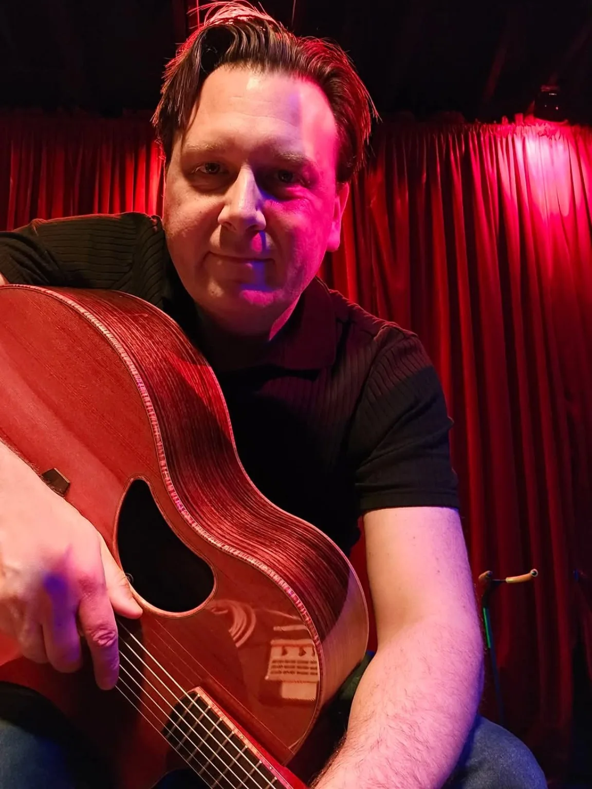 Dan Ennis with his guitar against a red stage curtain